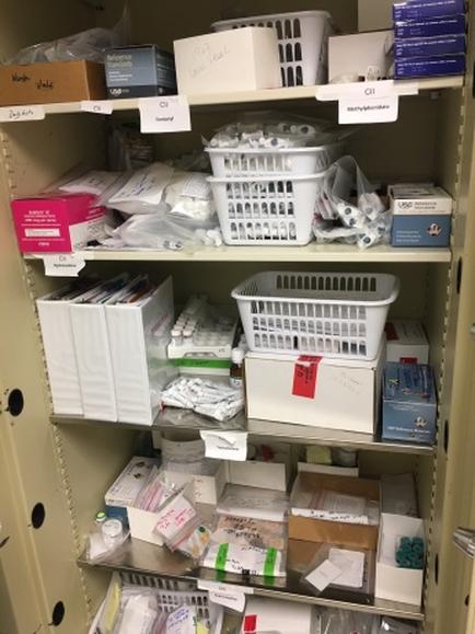 A metal cabinet with three shelves. The shelves are filled with various items such as boxes baskets and other medical supplies. The items are neatly arranged and appear to be of different sizes and colors. On the top shelf there is a white plastic basket with a label that reads "C1". On the bottom shelf on the left side there are several white boxes with labels that read "C2" "C3" and "C4". On top of the boxes there appears to be a pink box with a white label that says "C5". On both sides of the cabinet there seems to be more boxes and containers. The cabinet is open revealing the contents inside.