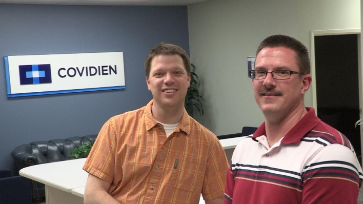 Two men standing in an office with a blue wall behind them. On the wall there is a sign that reads "COVIDIEN" in white letters. The man on the left is wearing an orange plaid shirt and is smiling at the camera. He has short blonde hair and is standing in front of a white desk with a black leather couch in the background. The other man is wearing a red and white striped polo shirt and has a mustache and glasses. Both men appear to be happy and relaxed.