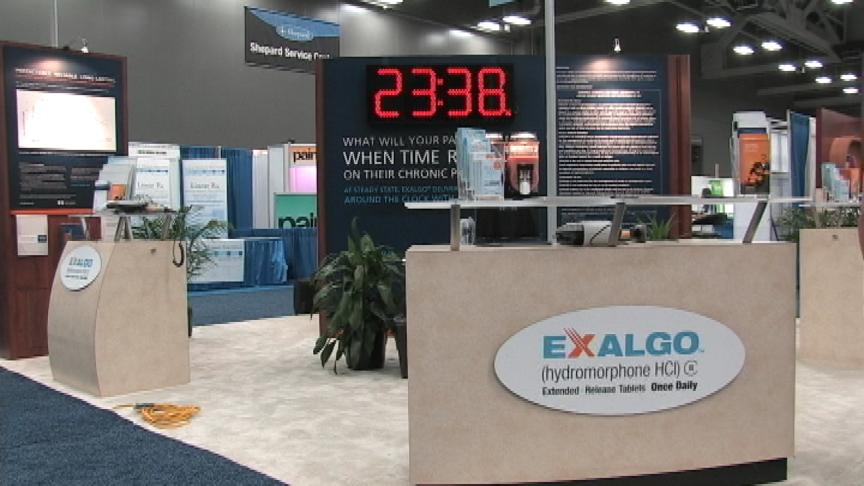 A trade show booth with a large digital display that reads "23:38". The booth is made of wood and has a beige color scheme. On the right side of the booth there is a desk with a computer monitor and other items on it. Behind the desk there are several other booths with information about the booth. The booth also has a sign that says "Exalgo (hydromorphone HCI) C" and a logo for Exalgo. The background of the image shows the floor and walls of the trade show.