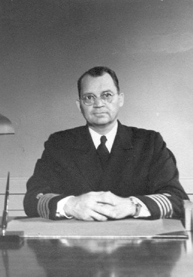 A black and white photograph of a man sitting at a desk. He is wearing a suit and tie and has a serious expression on his face. He has short dark hair and is wearing glasses. The desk is made of wood and there is a lamp on the left side of the image. The man is sitting with his hands clasped in front of him and appears to be deep in thought. The background is plain and the lighting is dim.