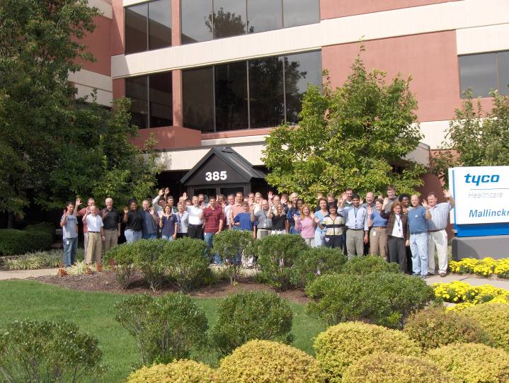 A group of people standing in front of a building with a sign that reads "tyco Mallinckrodt". The building appears to be a modern office building with large windows and a large entrance. The group is made up of men and women of different ages and ethnicities and they are all smiling and posing for a group photo. The building is surrounded by a well-manicured garden with shrubs and trees. The sky is blue and there are a few clouds in the background.