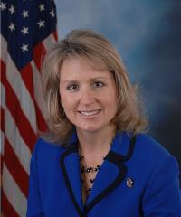 A portrait of a woman standing in front of an American flag. The woman is wearing a blue blazer with a black collar and a black necklace. She has shoulder-length blonde hair and is smiling at the camera. The background is blurred but it appears to be a dark blue color with a hint of smoke or fog. The American flag is visible on the left side of the image.