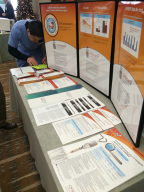 A table with three orange and white posters on it. The posters have text and images on them and there are several pages of information on the table. The table is covered with a gray tablecloth and there is a Christmas tree in the background. A man in a blue shirt is standing next to the table and appears to be working on one of the posters. He is looking at the posters with a focused expression on his face.