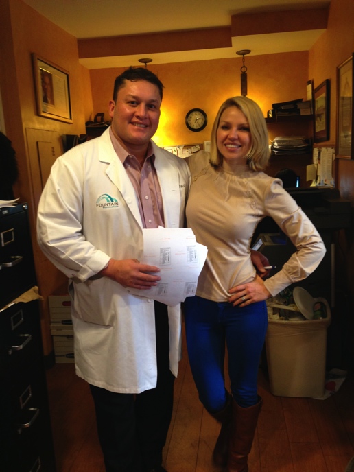 A man and a woman standing in a room with orange walls and wooden flooring. The man is wearing a white lab coat with a green logo on the left side and is holding a piece of paper in his left hand. He is smiling at the camera and the woman is standing next to him with her right hand on his hip. They are both dressed in formal attire with the man wearing a pink shirt and black pants and the women wearing beige blouses and blue jeans. There is a desk with a computer and a trash can in the background.