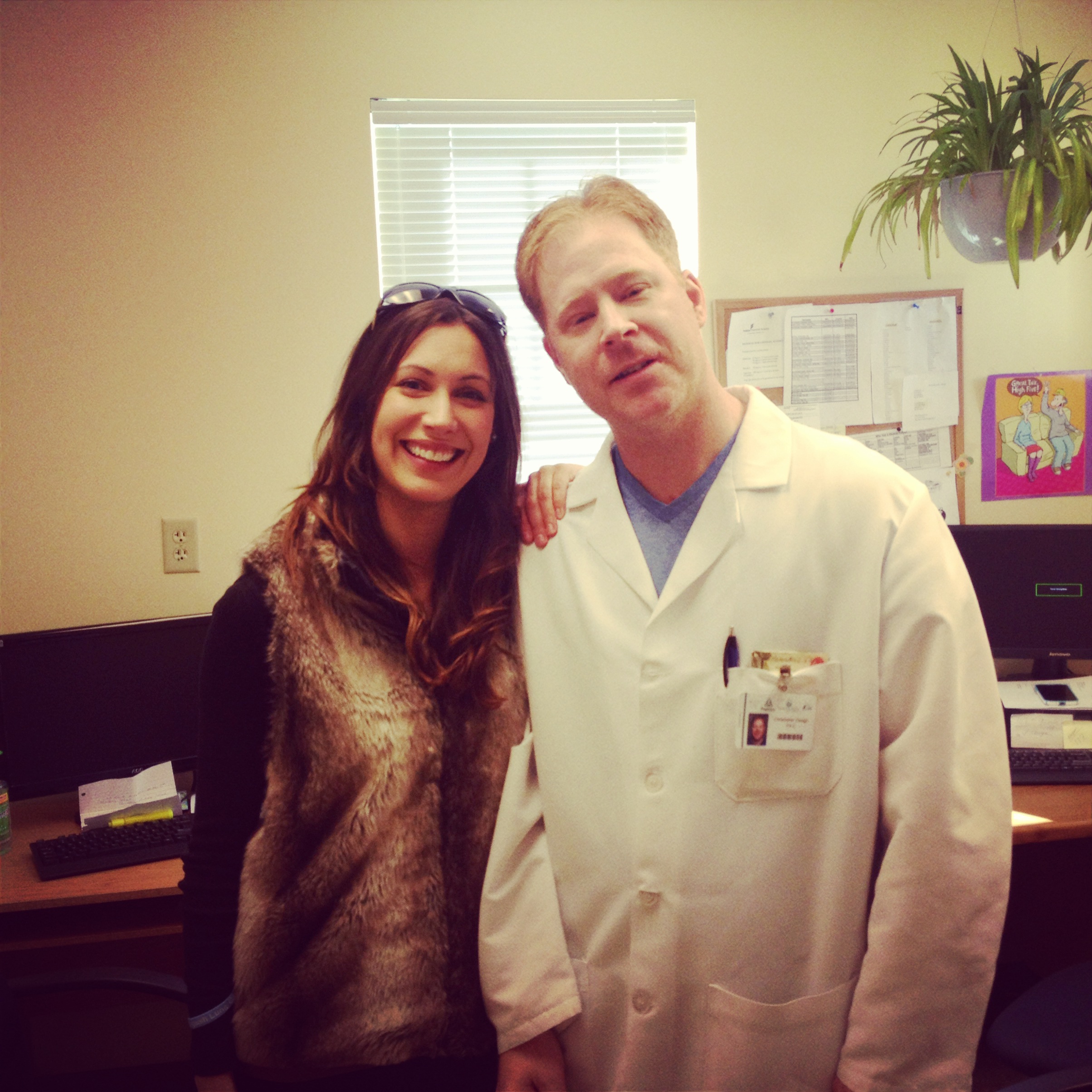 A man and a woman standing in an office. The man is wearing a white lab coat with a name tag and a pen in his pocket. He has short blonde hair and is smiling at the camera. The woman is also smiling and wearing a fur vest. They are standing next to each other and are standing in front of a desk with a computer monitor and other office supplies. There is a window with blinds in the background and a plant hanging from the ceiling.