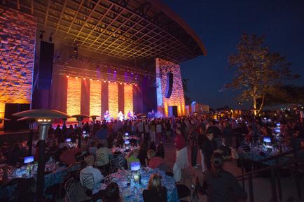 A large outdoor event space at night. The space is filled with people some sitting at tables and some standing. The tables are covered with white tablecloths and there are umbrellas providing shade. In the center of the space there is a stage with a band performing on it. The stage is decorated with colorful lights and there is an archway above it. On the right side of the image there are trees and buildings visible in the background. The sky is dark indicating that it is nighttime.