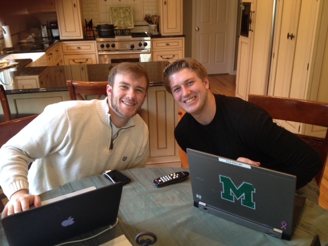 Two young men sitting at a kitchen table with two laptops in front of them. They are both smiling and appear to be happy. The man on the left is wearing a white sweater and is sitting on a wooden chair while the man in the black shirt is sitting next to him. Both men are looking at the camera and smiling. On the table there are two black laptops one with the letter "M" on the screen and the other with the logo of the University of Michigan on the lid. There is also a remote control on the table. The kitchen has wooden cabinets and a kitchen island in the background.