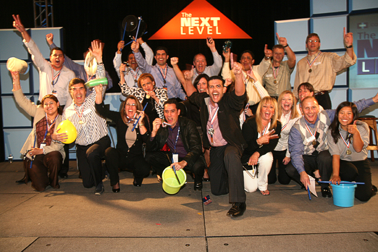 A group of people on a stage with their hands up in the air. They are all smiling and appear to be celebrating. In the center of the image there is a large orange triangle with the words "The Next Level" written on it. The people on the stage are dressed in formal business attire with some wearing suits and others wearing ties. Some of them are holding hard hats while others are holding buckets. The background is dark and there are banners on either side of the stage.