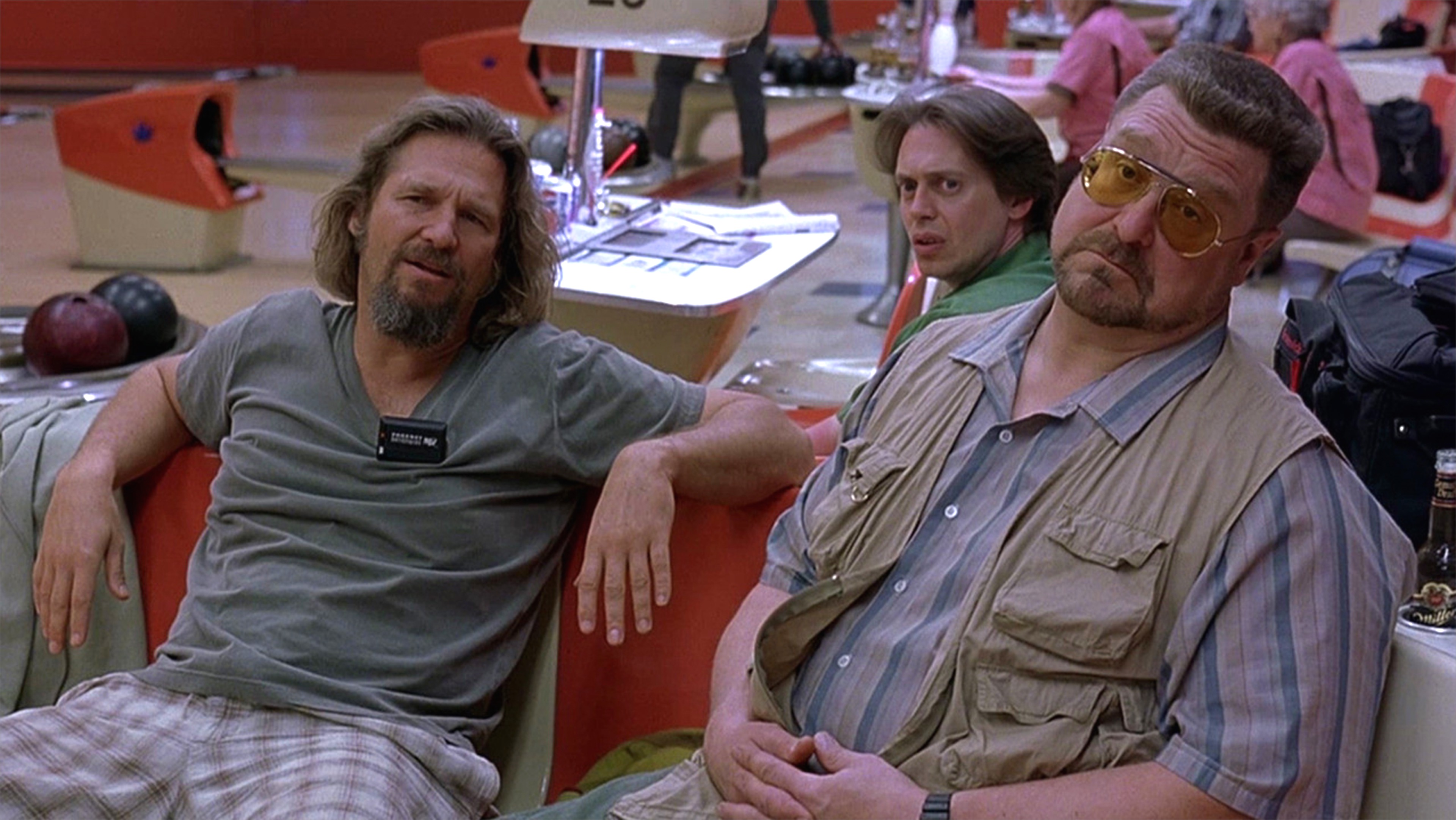 Three men sitting in a bowling alley. The man on the left is wearing a gray t-shirt and has long blonde hair and a beard. He is sitting on a red armchair with his legs crossed and is looking off to the side with a serious expression on his face. The other two men are sitting next to him one wearing a green shirt and the other wearing a beige shirt and sunglasses. They are all looking at the camera and appear to be deep in thought. In the background there are several bowling pins and other people in the lanes.