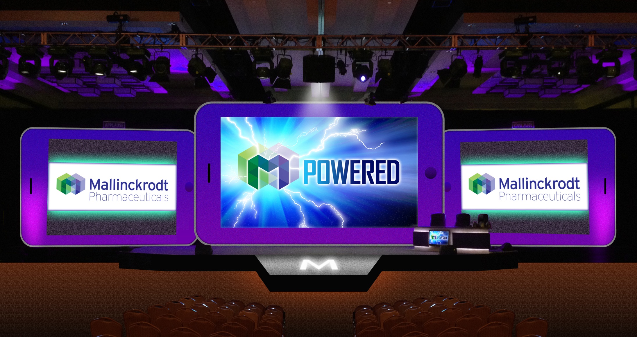 A stage set up for a presentation. There are three large screens on the stage each displaying the logo of Mallinckrodt Pharmaceuticals. The screens are arranged in a triangular formation with the largest screen in the center and two smaller screens on either side. The background of the stage is black and there are rows of empty seats in front of it. The stage is lit up with purple and blue lights creating a dramatic atmosphere.