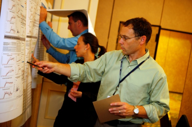 A group of three people in a room with a large whiteboard on the wall. The whiteboard is filled with graphs and charts that appear to be related to a presentation or presentation. The man in the foreground is pointing to one of the charts on the board while the other two people are looking at it intently. He is wearing a light green shirt and glasses and is holding a brown folder in his hand. The woman in the middle is standing next to him and the man on the left is leaning over the board and appears to be explaining something to the group. The background is a beige wall with a wooden panel on the right side. It appears that the group is in a conference room or a meeting room.