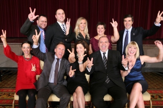 A group of ten people posing for a photo on a stage with a red curtain in the background. All are dressed in formal attire. They are all smiling and making a peace sign with their hands. The men are wearing suits and ties while the women are wearing dresses. The group appears to be happy and confident and the overall mood of the photo is joyful and celebratory.