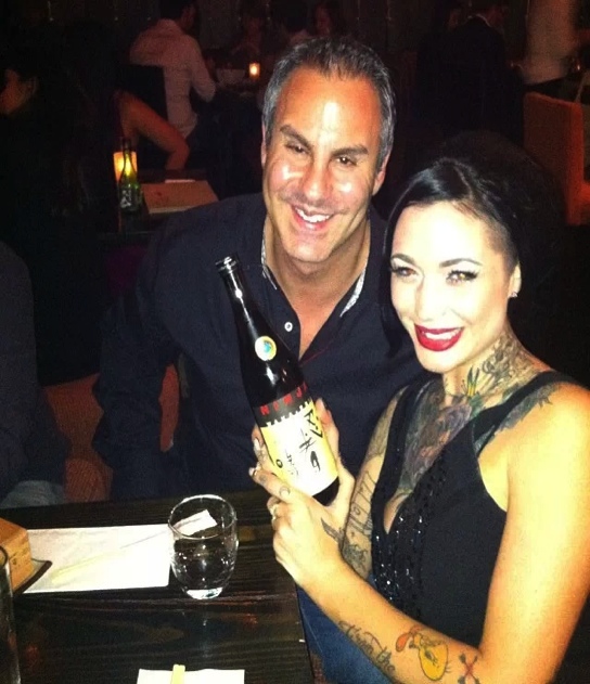 A man and a woman sitting at a table in a restaurant. The man is holding a bottle of champagne and the woman is smiling at the camera. They are both dressed in formal attire with the man wearing a black shirt and the women wearing black dresses. The woman has tattoos on her arms and neck and she has dark hair and red lipstick. There is a glass of water on the table in front of them. The background is blurred but it appears to be a dimly lit restaurant with other people sitting at tables.