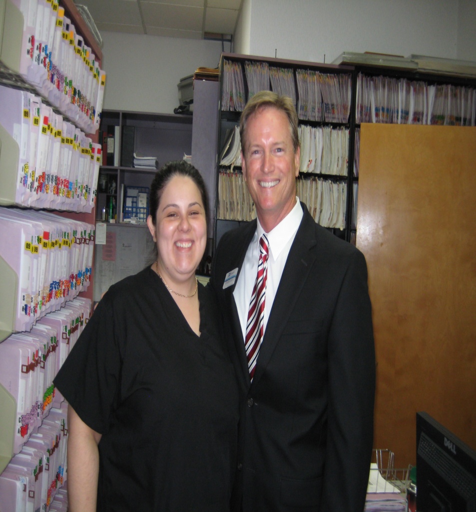 A man and a woman standing in an office. They are both smiling and posing for the camera. The man is wearing a black suit with a red and white striped tie and a name tag on his lapel. He has blonde hair and is standing next to the woman. Behind them there are shelves filled with folders and binders. On the right side of the image there is a desk with a computer monitor and other office supplies. The office appears to be well-lit with natural light coming in from the windows.