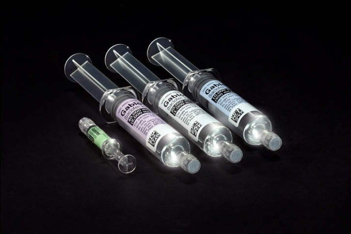 Four syringes of different sizes and shapes arranged in a row on a black background. The syringes are made of clear plastic and have a cylindrical shape with a pointed tip. Each syringe has a label on it with a white background and black text. The label also has a barcode and some other information printed on it. There is also a small green syringe lying next to the syringes. The overall color scheme of the image is black and white.