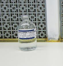 A small glass bottle with a white cap and a blue label. The label has the text "BACLOFEN S.O.D.M.G.M." written on it in white letters. The bottle appears to be made of clear glass and is sitting on a white surface. In the background there is a metal grille with a geometric pattern. The grille is slightly blurred but it seems to be placed against a white wall.