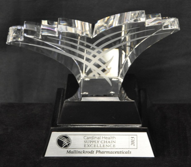 Of a crystal trophy on a black base. The trophy is made of clear glass and has a unique design with a curved shape that resembles a wave or a wave-like pattern. The base is rectangular in shape and has the words "Cardinal Health Supply Chain Excellence" and "Mallinckrodt Pharmaceuticals" engraved on it. The background is black making the trophy stand out.