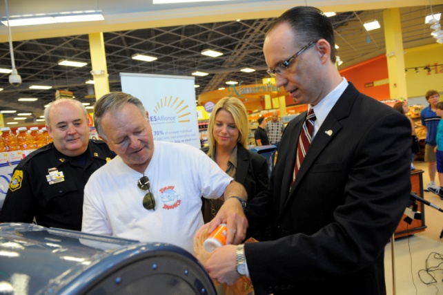 A group of people in a grocery store. There are four people in the foreground three men and a woman. Two of the men are holding a single bottle of pills and appear to be pouring them into a blue trash can. In the background there are shelves stocked with bottles of juice and other items. The store appears to have a high ceiling with overhead lights.