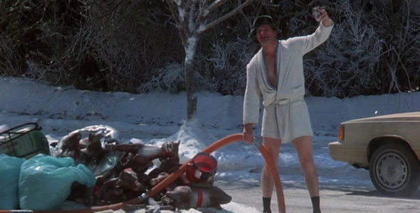 A man standing on a snowy road with his arms raised in the air. He is wearing a white robe and a green hat. He has a big smile on his face and appears to be happy. In front of him there is a pile of trash and debris including a red hose and a blue bag. There is also a car parked on the side of the road. The background shows trees and bushes covered in snow.