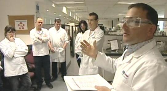 A group of six people in a laboratory setting. They are all wearing white lab coats and are standing in a line with their arms crossed. The person in the front is a man wearing a white lab coat and glasses and he is gesturing with his hands as he speaks. Behind him there are shelves with various scientific equipment and a bulletin board with a picture of a person on it. The group appears to be engaged in a discussion or presentation.