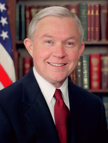 A portrait of a middle-aged man in a formal setting. He is wearing a dark suit and a red tie. He has short white hair and is smiling at the camera. Behind him there is a bookshelf filled with books and an American flag. The man appears to be in his late 60s or early 70s.