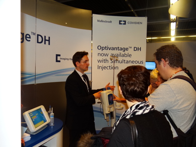 A man in a suit standing in front of a booth at a trade show. He is explaining something to a group of people who are gathered around him. The booth has a banner that reads "Optivantage™ DH now available with Simultaneous Injection" and there is a computer monitor on the table next to the booth. The man appears to be explaining the treatment to the people in the group.