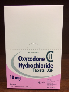 A photograph of a white box of Oxycodone Hydrochloride tablets. The box is rectangular in shape and has a pink label on the front. The label has the brand name "Oxycodone" written in bold black letters at the top followed by the product name "Hydrochloride Tablets USP" in smaller black letters. Below that there is a description of the product which states that it is 10mg in size and contains 100 tablets. At the bottom of the label there are two small logos one for "Rhodes" and the other for "10mg". The box appears to be sitting on a wooden surface.