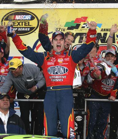 A race car driver Dale Earnhardt Jr. standing on the podium with his arms raised in triumph. He is wearing a red and blue racing suit with the words "Outpoint Automotive Finishes" written across the chest. He has a big smile on his face and is holding a bottle of champagne in his right hand. In the background there are several other race car drivers and a large banner that reads "NASCAR Nextel Cup Series". The image appears to have been taken at a NASCAR event with confetti and streamers in the air.