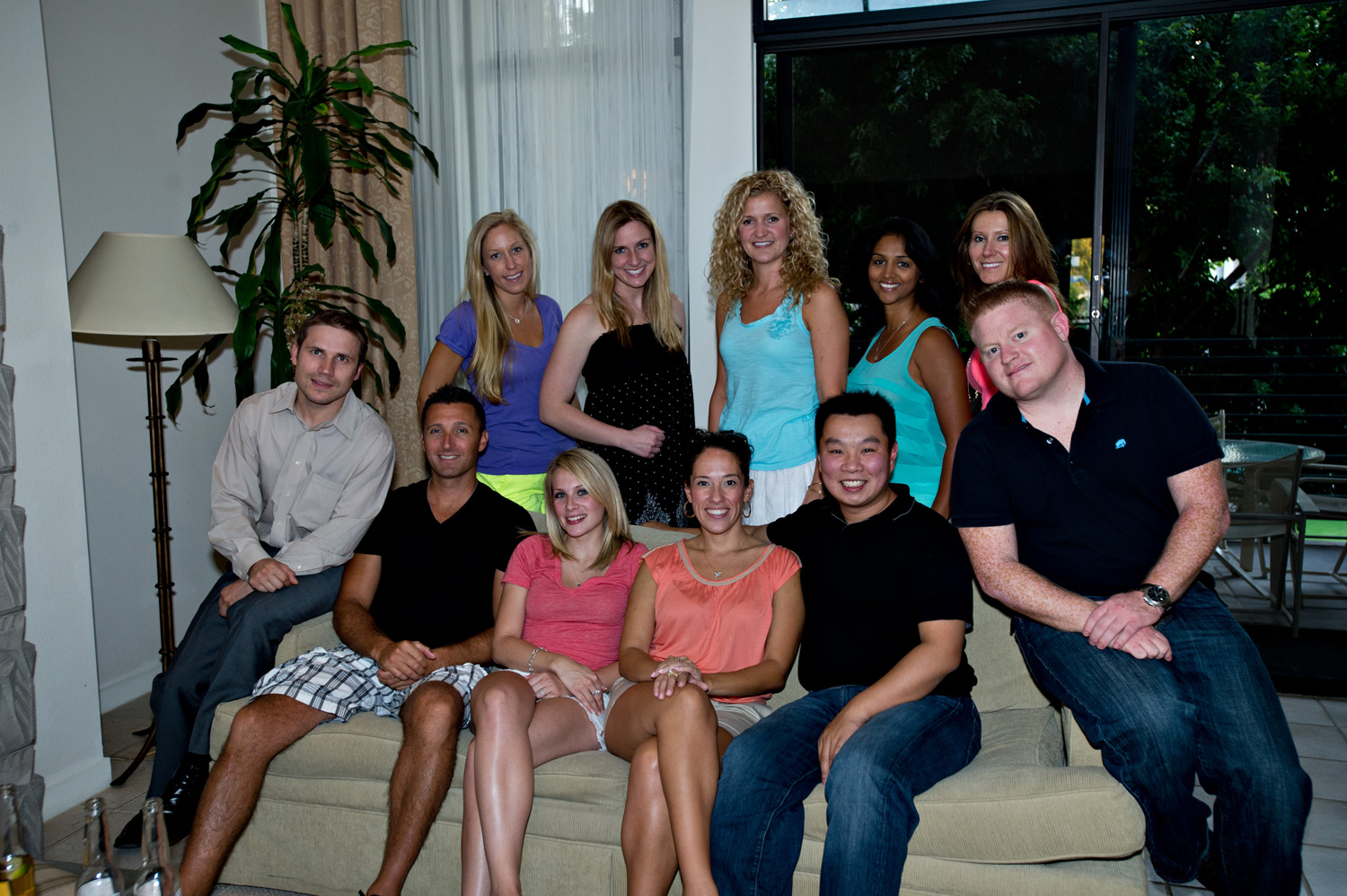 A group of eleven people sitting on a beige couch in a living room. All are smiling and posing for the camera. They are all wearing casual clothes and appear to be of different ages and ethnicities.<br /><br />In the background there is a large window with white curtains and a potted plant on the windowsill. The room is decorated with a gray rug and a lamp on the floor. There is also a dining table and chairs visible in the background. The overall mood of the image is happy and relaxed.