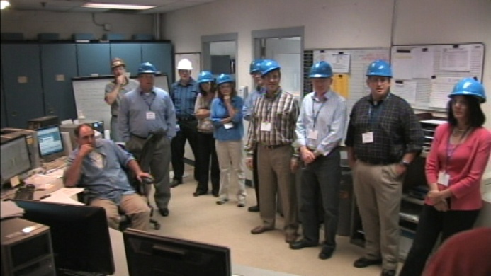 A group of people in an office setting. There are nine people in the group all wearing blue hard hats. They are standing in front of a desk with multiple computer monitors and keyboards. On the left side of the image there is a man sitting on a chair with his hand on his chin looking at the camera. He appears to be deep in thought. In the background there are lockers and a bulletin board with papers pinned to it. The overall atmosphere of the room is professional and organized.