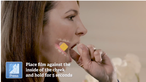 A close-up of a woman's face with her mouth slightly open and her hand resting on her chin. She appears to be deep in thought with a yellow and pink checkmark on her cheek. The background is blurred but it seems like she is in a room with white flowers. The text on the image reads "Place film against the inside of the cheek and hold for 5 seconds".