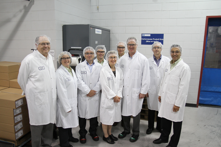 A group of people standing in a large room with white walls and a blue door on the right side. They are all wearing white lab coats and head caps and are smiling at the camera. In the background there are stacks of cardboard boxes and a large machine. The group appears to be in a laboratory or workshop as there is a sign on the wall behind them that reads "Certificate of Authenticity". It seems like they are posing for a group photo.