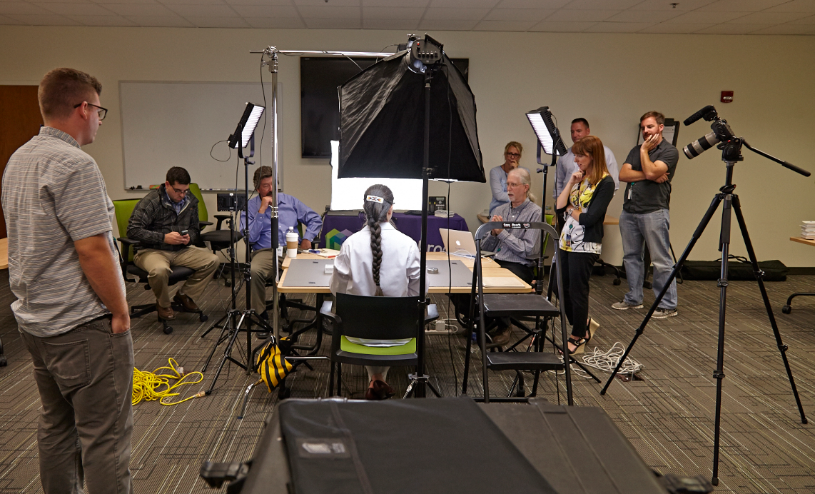 A group of people in a room with a large camera setup. There are six people in the room three men and three women all of whom appear to be engaged in a conversation. The room has a carpeted floor and a whiteboard on the wall in the background.<br /><br />In the center of the image there is a large black camera with a tripod stand in front of it. The camera is set up on a tripod and there are several other cameras and equipment scattered around the room. On the left side of the photo there are two men sitting at a desk one of whom is holding a camera and the other is looking at the camera. The other two men are standing on the right side and the third man is standing in the center holding a microphone and appears to be speaking to the group. There is also a woman sitting on a chair in the foreground who is wearing a white shirt and has her back to the camera and another man standing next to her looking at her with a serious expression on his face.