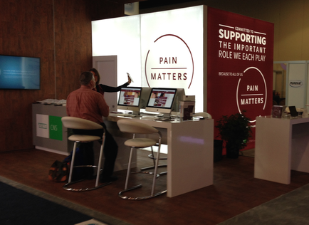 A booth with a red and white banner that reads "Pain Matters" and "Supporting the important role we each play". The booth is located in a room with wooden walls and a wooden floor. There are two people sitting at a desk in front of the desk one of them is a man and the other is a woman. They are both looking at a computer screen and appear to be engaged in a conversation. On the desk there are two computer monitors and a coffee cup. There is also a plant in the corner of the booth.
