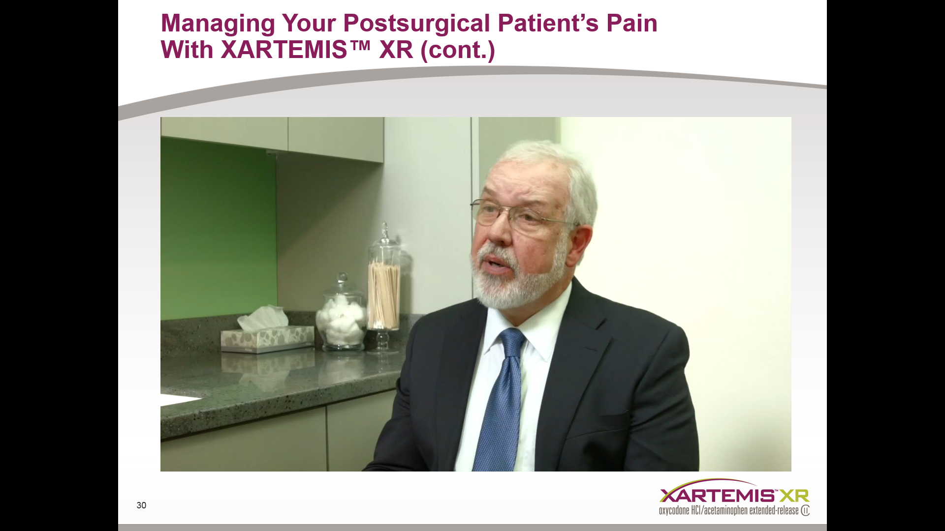 A screenshot from a video titled "Managing Your Postsurgical Patient's Pain With Xartemis™ XR (cont.)". It shows a man in a suit and tie sitting in a room with a green wall and a countertop in the background. He appears to be in the middle of a conversation with a serious expression on his face. The title of the video is written in white text at the top of the image.