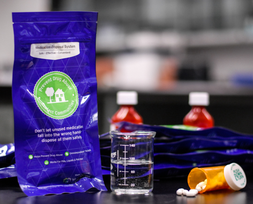 A blue packet of medication disposal system on a black countertop. The packet has a green logo and text on it that reads "Prevent Drug Abuse". Next to the packet there is a beaker filled with water and a small orange pill bottle with white pills spilling out of it. There are also two small bottles of red liquid one with a white cap and the other with a red cap. The background is blurred but it appears to be a kitchen or laboratory setting.