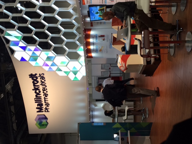 A trade show booth with a modern design. The booth has a white background with a colorful geometric pattern on the left side. On the right side there is a large screen displaying information about the booth. In the center of the booth there are several tables and chairs arranged in a semi-circle. A man is standing in front of one of the tables working on a laptop. There are other people in the background and the booth appears to be well-lit.