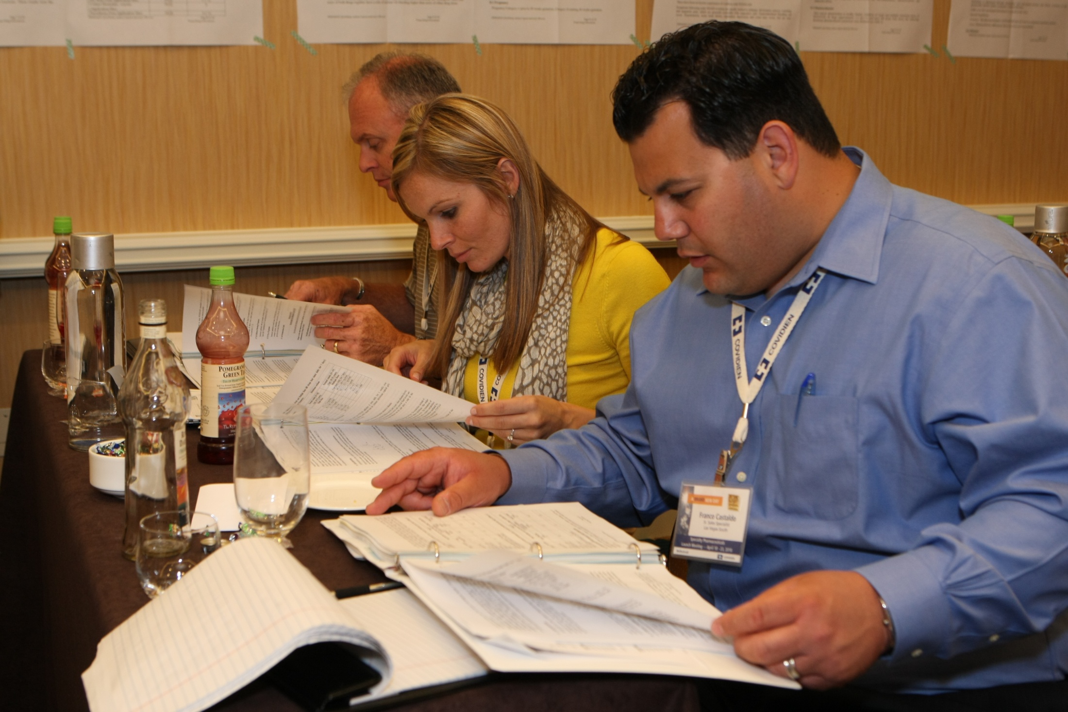 Three people sitting at a long table in a conference room. The table is covered with a brown tablecloth and there are several bottles and glasses on it. The person on the left is a man the person in the middle is a woman and the man on the right is a young man. They are all looking at a piece of paper and appear to be working together on a project. The woman is wearing a yellow blouse and the two men are wearing blue shirts. All three people are focused on their work and seem to be engaged in the task at hand.