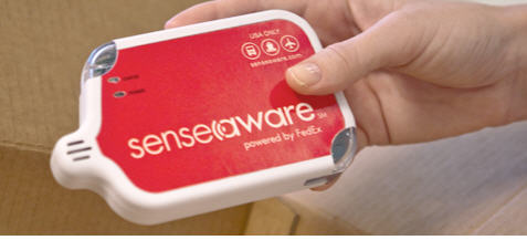 A hand holding a red and white device with the word "senseaware" written on it. The device appears to be a wireless device as indicated by the logo on the top left corner of the device. The logo is a red circle with a white "S" in the center and the words "powered by FedEx" written in white below it. On the top right corner there is a small icon of a person's hand holding the device which is likely a logo for the company. The background is blurred but it seems like the device is placed on a wooden surface.