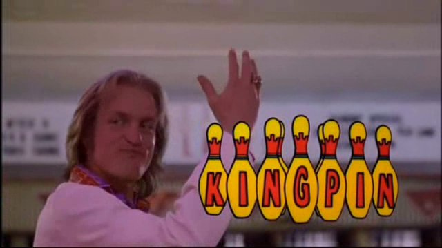 A still from a movie or TV show. It shows a man with long blonde hair and a purple scarf around his neck standing in front of a bowling alley. He is smiling and waving to the camera with his right hand. In front of him there are six bowling pins arranged in a row with the word "KINGPIN" written above them in red and yellow letters. The background is blurred but it appears to be an indoor bowling alley with other lanes visible.