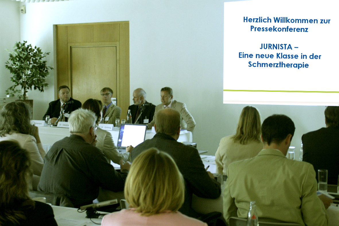 A group of people sitting at a long table in a conference room. The room has white walls and a wooden door in the background. There is a projector screen on the right side of the image with the text "Herzlich Willkommen zur Pressekonferenz Jurnista - Eine neue Klasse in der Schmerztherapie" which translates to "The presentation of the new Klasse of the Schmerzertherapies" in English. The people in the image appear to be engaged in a discussion with some sitting at the table and others standing at the front of the room. They are all dressed in formal business attire and are looking towards the projector screen.