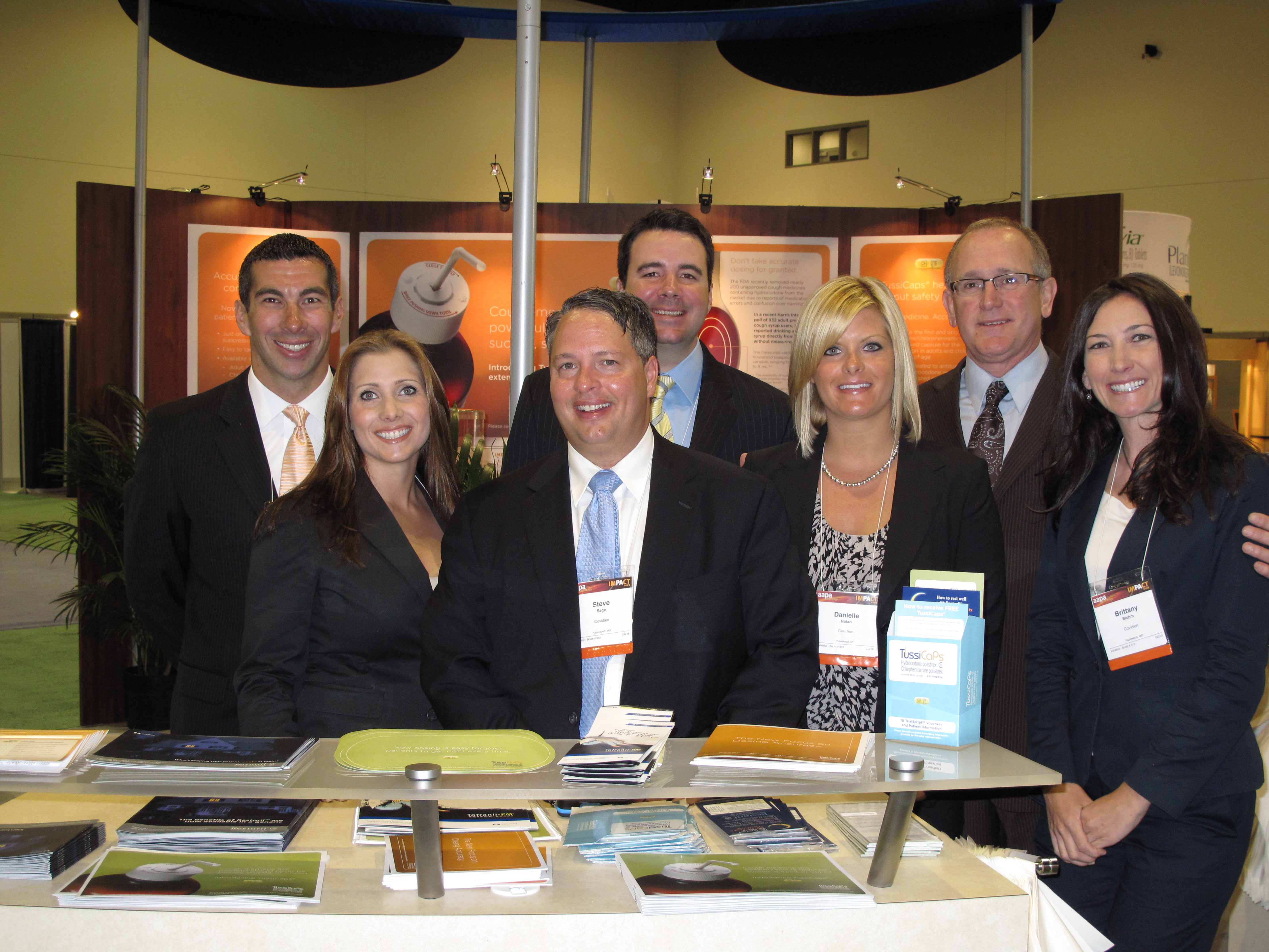 A group of seven people standing behind a table with books on it. They are all dressed in formal business attire and are smiling at the camera. The table is covered with a white tablecloth and there are several books on the table. In the background there is a banner with an orange and white design and a blue canopy. The people appear to be at an event or trade show.