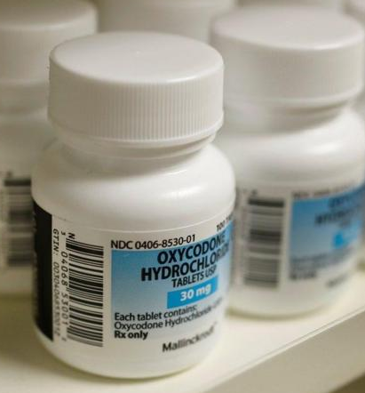 A row of white medicine bottles with white caps. The bottles are arranged in a row on a white shelf. Each bottle has a blue label with the text "NDC 0406-8530-01 Oxycodone Hydrochloride Tablets" and a barcode. The label also mentions that each tablet contains 30mg of Oxycodone Hydrochloride Rx only. The background is blurred but it appears to be a pharmacy or drugstore.