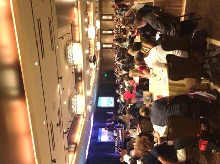 A long hallway with a group of people sitting at tables. The hallway is dimly lit with chandeliers hanging from the ceiling. On the left side of the hallway there is a white door with a chandelier hanging above it. The people are sitting on chairs and appear to be engaged in conversation. In the background there are stairs leading up to a stage with a blue curtain. The overall mood of the image is casual and relaxed.