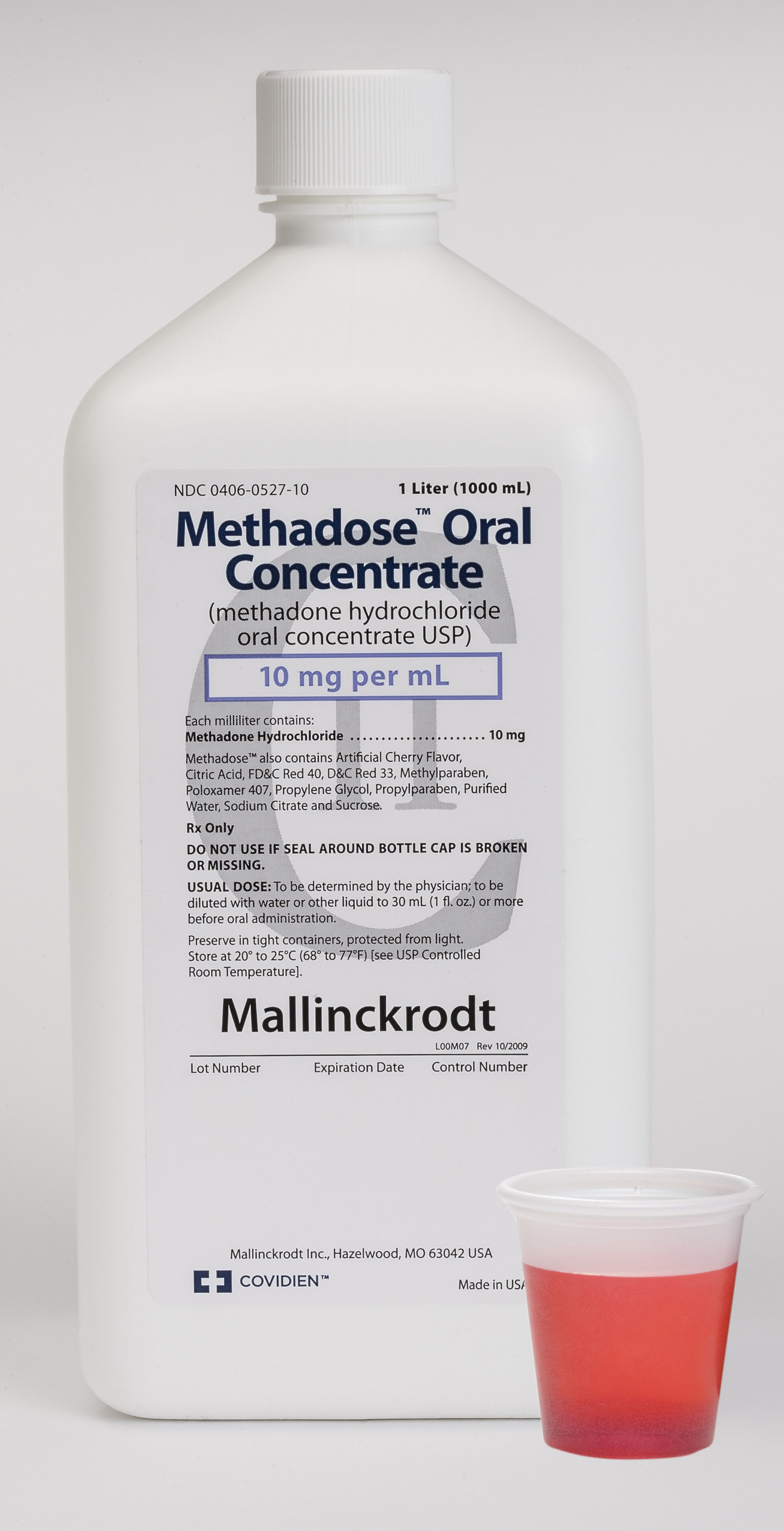 A white plastic bottle with a white cap and a red plastic cup next to it. The bottle is labeled "Methadose Oral Concentrate" and has a label that reads "10 mg per ml". The label also mentions that the product is made in the USA and is called "Mallinckrodt". The bottle appears to be new and unused. The cup is empty and is placed next to the bottle. The background is plain white.