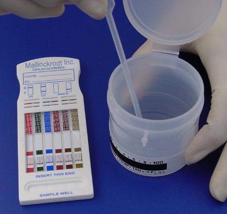 A hand wearing a white glove holding a small plastic container with a lid. The container has a label on it that reads "Mallinckrodt Inc." and there is a test strip next to it. There are multiple rows of test strips with different colors and labels. The label also has the words "Insert This End" and "Sample Well" written on it indicating that the test strip is being inserted into the container. The background is a plain blue surface.