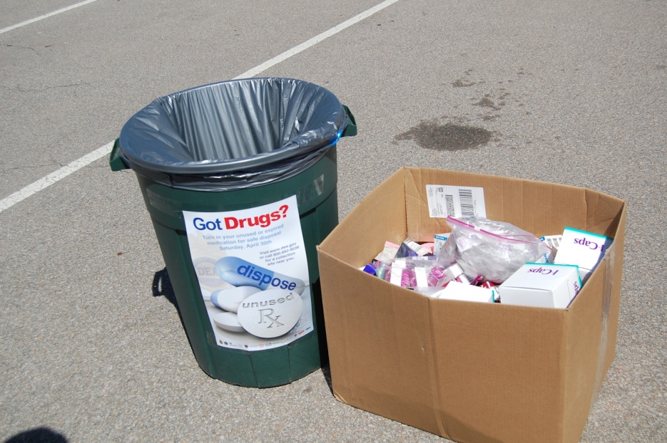 A green trash can with a label that reads "Got Drugs?" next to a cardboard box filled with various items. The trash can appears to be empty and the cardboard box is overflowing with items such as pills capsules and other medical supplies. The background shows a parking lot with white lines marking the lanes.