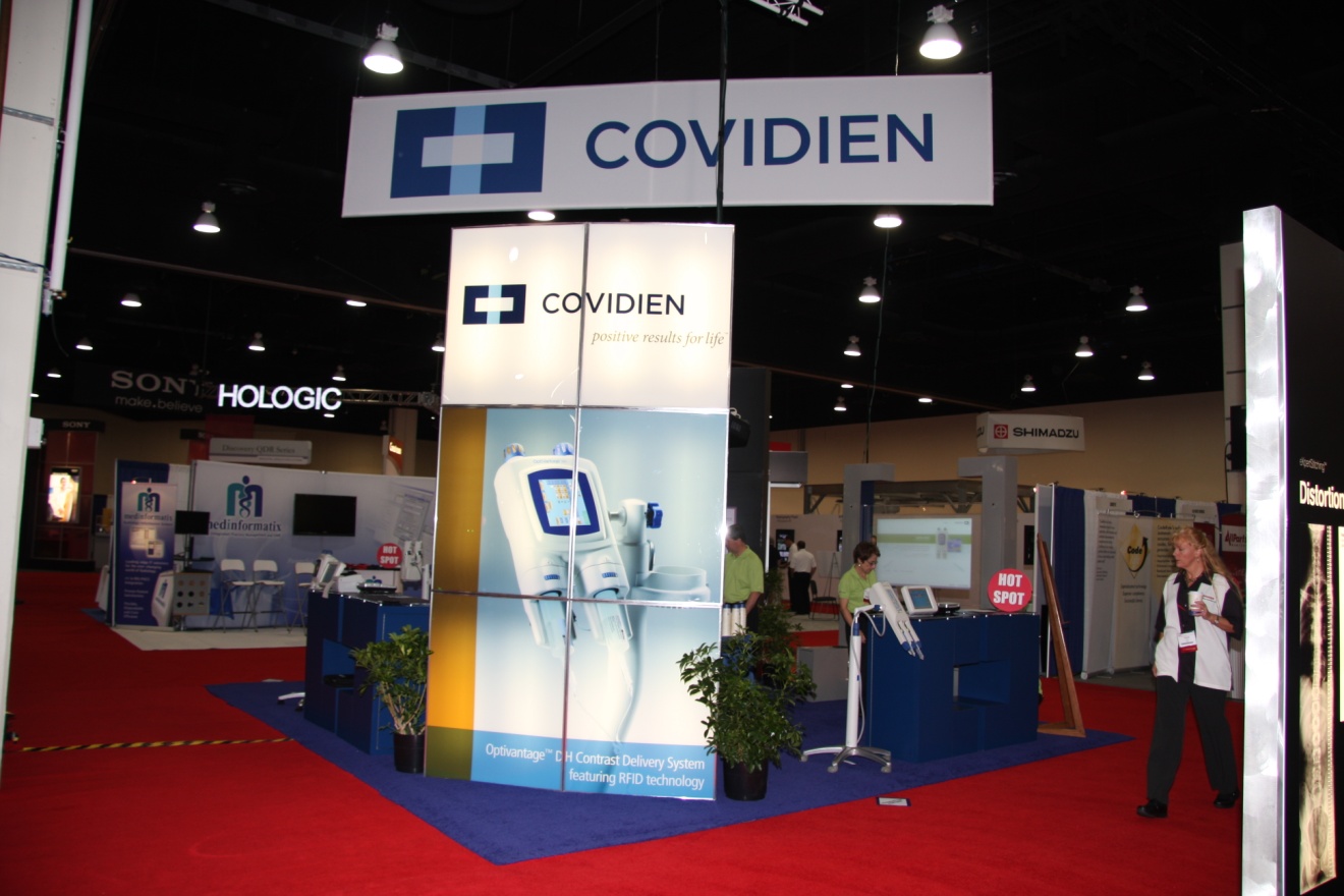 A trade show booth with a blue and white color scheme. The booth has a large banner that reads "COVIDIEN" in bold letters at the top. Below the banner there is an image of a medical device which appears to be a CT scan machine displayed on a blue background. On the right side of the booth there are several other booths with information about the product such as "SONY HOLOGIC" and "Covidien". There are also several people standing in front of the booths looking at the display. The floor is covered in a red carpet and there are potted plants scattered around the booth.