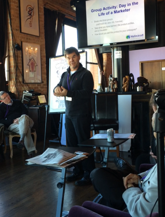 A man standing in front of a projector screen in a room with brick walls and wooden flooring. He is holding a microphone and appears to be giving a presentation. There are three people sitting in chairs in the room one of whom is holding up a camera and taking a picture of the man. On the right side of the image there is a table with a newspaper and a can of soda on it. The room is decorated with various artworks and a large window with curtains. The man is wearing a black jacket and has a serious expression on his face.