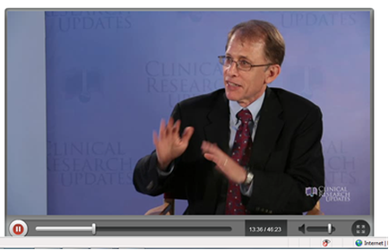A screenshot of a video clip of a man in a suit and tie sitting on a chair in front of a blue background with the words "Clinical Research Updates" written on it. The man appears to be in the middle of a presentation as he is gesturing with his hands as if he is explaining something. He is wearing glasses and has a serious expression on his face. The date and time of the video is 13:36:45:23.